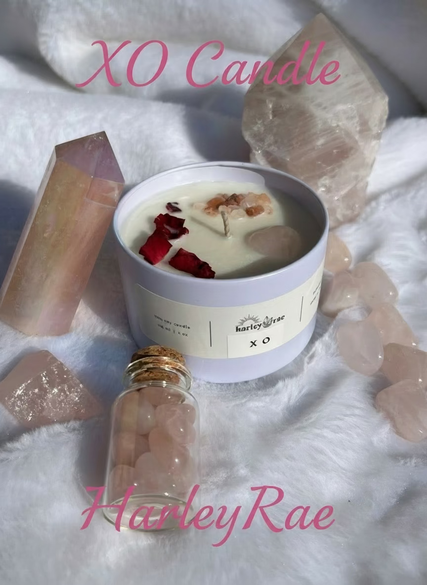 A soy candle with a blend of aromatic herbs, warm amber, and florals, topped with dried rose petals, Himalayan pink salt, and a Rose Quartz heart, presented in a white jar surrounded by decorative stones and a small glass bottle.