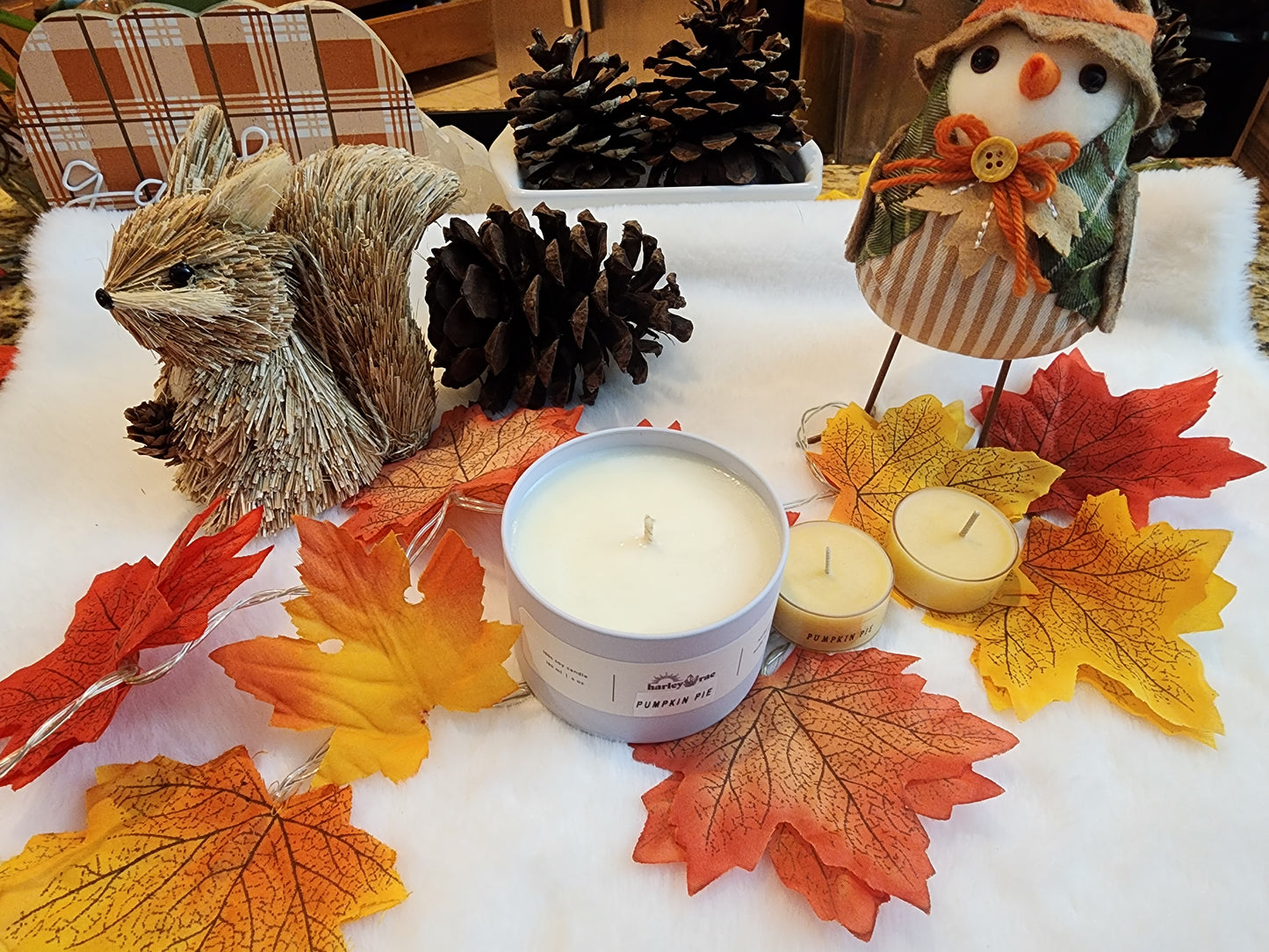 A beige candle with a pumpkin pie scent, displayed on a white surface surrounded by autumn-themed decorations including leaves, a squirrel, and a snowman.