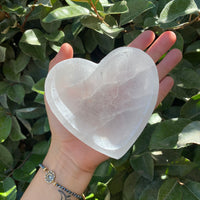 A person's hand holding a heart-shaped selenite crystal bowl with a background of green leaves.