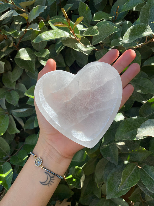 A person's hand holding a heart-shaped selenite crystal bowl with a background of green leaves.