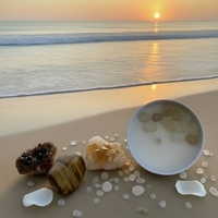 A white candle with clear liquid and citrine crystals on top, placed in a white bowl, with a wooden block and loose citrine crystals to the side, all set against a white fuzzy background.