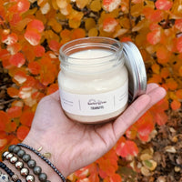 Thankful jar candle with a white substance inside, in front of a background with autumn-colored leaves.